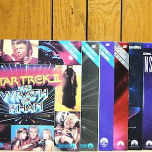 Può includere: Una collezione di videocassette VHS e LaserDisc vintage di Star Trek II: L'ira di Khan. Le copertine presentano immagini del cast e scene del film, con colori vivaci e il titolo del film. Include versioni Extended Play.