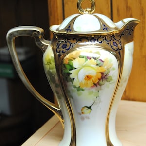Puede incluir: Una tetera de porcelana antigua con tapa, con un diseño floral pintado a mano. La tetera tiene una base blanca con ribetes dorados y un asa curvada. El diseño floral incluye rosas amarillas y naranjas.