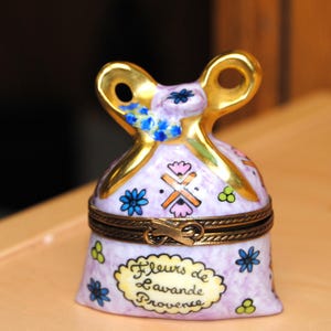 Könnte beinhalten: Eine lavendelfarbene Keramik-Schmuckdose in Form eines Beutels, mit goldenen Akzenten und einem Klappdeckel. Verziert mit floralen Motiven und dem Text "Fleurs de Lavande Provence".