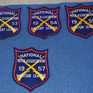 May include: Four blue and red embroidered patches with the text "National Rifle Association" and crossed rifles. The patches have different years and league names: "19 53 Gallery League", "19 54 Gallery League", "19 55 Winter League", and "19 57 Winter League".