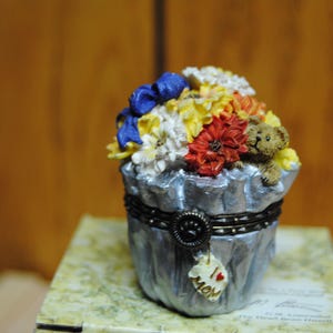 以下が含まれることがあります： 色とりどりの造花と小さなテディベアが入った、花瓶型の装飾的な小物入れ。ボックスはシルバーで、濃い色の金属バンドと「I Love Mom」と書かれたチャームが付いています。