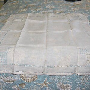 May include: White linen tablecloth with delicate embroidered details. The tablecloth has a square shape and a scalloped edge. It is folded in half and laid on a blue and white patterned surface.