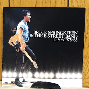 Puede incluir: Portada del álbum "Live/1975-85" de Bruce Springsteen & The E Street Band. La imagen presenta una foto de un concierto de un hombre sosteniendo una guitarra, con el nombre de la banda y el título del álbum en texto blanco sobre fondo negro.