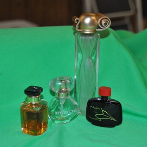 May include: Four different perfume bottles on a green surface. The bottles are all different shapes and sizes. The bottle on the left is a clear glass bottle with a gold cap. The bottle in the centre is a clear glass bottle with a gold cap and a black label that says "Intuition". The bottle on the right is a black glass bottle with a red cap and a gold label that says "Maxim's Paris".