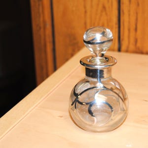 Puede incluir: Un frasco de perfume de vidrio transparente con un cuello y tapón de metal plateado. La botella tiene forma redonda con un diseño de remolino plateado. El tapón es de forma redonda y transparente. La botella está sobre una superficie de madera clara.