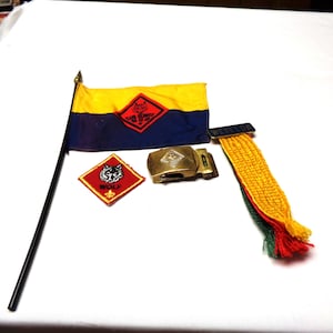 May include: A set of Boy Scout memorabilia including a yellow and blue flag with the Scout emblem, a red and black patch with a wolf, a gold belt buckle with the Scout emblem, and a yellow, red, and green sash.