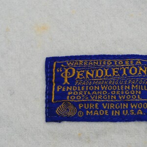 May include: A rectangular blue fabric clothing label with gold embroidered text that reads Warranted to be a Pendleton, Pendleton Woolen Mills, Portland, Oregon, 100% Virgin Wool, Pure Virgin Wool, and Made in U.S.A. with a yarn ball icon.