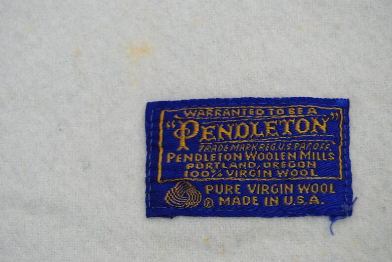 May include: A rectangular blue fabric clothing label with gold embroidered text that reads Warranted to be a Pendleton, Pendleton Woolen Mills, Portland, Oregon, 100% Virgin Wool, Pure Virgin Wool, and Made in U.S.A. with a yarn ball icon.