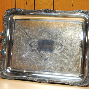Puede incluir: Bandeja rectangular plateada con detalles florales y de volutas ornamentadas. La bandeja presenta asas decorativas y una superficie reflectante. Una pieza vintage para servir o exhibir.
