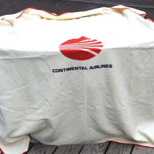 May include: An off-white blanket featuring the red Continental Airlines logo and the text CONTINENTAL AIRLINES in dark blue lettering. The blanket has a thin red border along the edges and a soft, woven texture throughout the fabric surface.