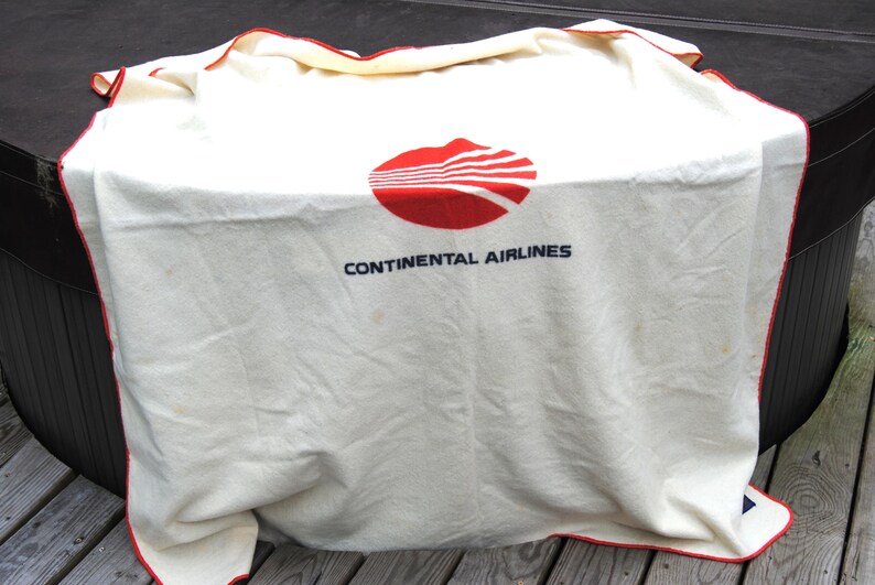 May include: An off-white blanket featuring the red Continental Airlines logo and the text CONTINENTAL AIRLINES in dark blue lettering. The blanket has a thin red border along the edges and a soft, woven texture throughout the fabric surface.