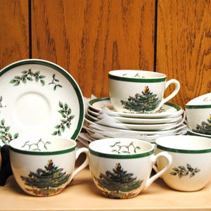 Könnte beinhalten: Ein Set aus weißen Keramik-Teetassen und Untertassen mit grünem Rand und Weihnachtsbaum-Design. Die Untertassen und Tassen sind gestapelt und auf einer Holzoberfläche angeordnet. Das Set enthält sechs Tassen und Untertassen.