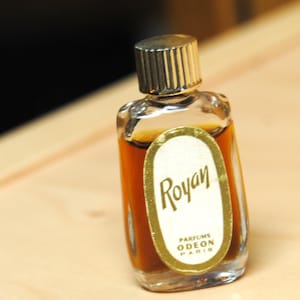 May include: A small, vintage perfume bottle with a gold-colored cap and a label that reads "Royan, Parfums Odeon Paris." The bottle is filled with a dark amber liquid and is set against a light-colored background.