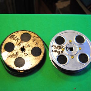 Puede incluir: Dos carretes de película vintage uno junto al otro sobre una superficie verde. Un carrete es dorado con círculos negros y texto, el otro es plateado con círculos negros y texto. El texto de los carretes es parcialmente visible.
