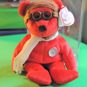 Puede incluir: Un osito de peluche rojo con un gorro de aviador marrón y gafas. El oso lleva una bufanda blanca y tiene una etiqueta Ty roja y blanca unida a su oreja. El oso está sentado sobre una superficie verde.