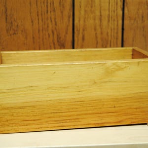 Puede incluir: Una caja rectangular de madera con la parte superior abierta. La caja está hecha de madera de color claro con vetas visibles y un acabado natural. La caja está sobre una superficie de color claro.