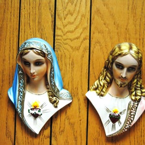 May include: Two religious wall hangings. One depicts a woman with a blue veil, the other a man with long, golden hair. Both have a white garment with a heart and flame design. The background is a wooden surface.