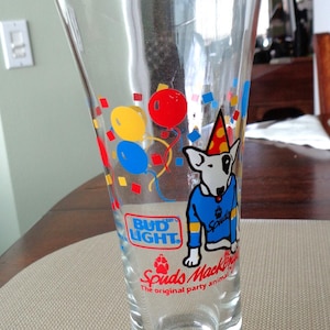 May include: Clear glass Bud Light beer glass with a wide base and flared top. The glass features a cartoon dog wearing a party hat and a blue shirt. Colorful balloons and the text "Bud Light" are also visible.