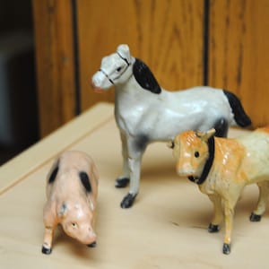 Puede incluir: Un conjunto de figuras en miniatura de animales de granja. El conjunto incluye un caballo blanco con crin negra, un cerdo rosa con manchas negras y una vaca marrón y blanca. Las figuras están dispuestas sobre una superficie clara.