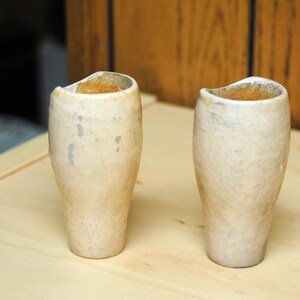May include: Two off-white, cylindrical vases with a curved top edge are displayed on a light wooden surface. The vases have a textured appearance with subtle gray markings. The background features a wooden wall.