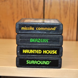 Może przedstawiać: Stos czterech czarnych kartridży do gier wideo Atari. Na górnym kartridżu napis "missile command" w żółtym tekście. Poniżej znajdują się kartridże z napisami "BERZERK", "HAUNTED HOUSE" i "SURROUND" w zielonym i żółtym tekście.