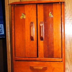 May include: A small, wooden cabinet with a warm, reddish-brown finish. The cabinet features two doors with vertical wood grain and two small decorative stickers. It has a single drawer at the bottom and two wooden handles. The cabinet is approximately 18 inches tall.