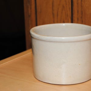 Può includere: Un vaso di gres cilindrico beige con un bordo arrotondato. Il contenitore è appoggiato su una superficie di legno chiaro, con uno sfondo scuro e una parete di legno sullo sfondo.