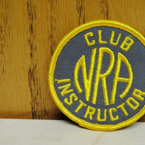 Op de afbeelding: Ronde patch met een grijze achtergrond en gele geborduurde tekst en rand. De tekst luidt "CLUB NRA INSTRUCTOR". De patch ligt op een lichtgekleurd oppervlak met een houten achtergrond.