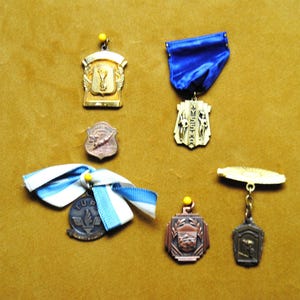 May include: A collection of vintage medals and pins in various shapes and colors. The medals include gold, bronze, and silver tones, some with ribbons. The pins and medals are pinned to a tan surface.