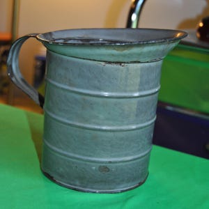 Puede incluir: Una jarra de esmalte gris claro vintage con asa. La jarra tiene un aspecto ligeramente desgastado y rústico y está hecha de metal.