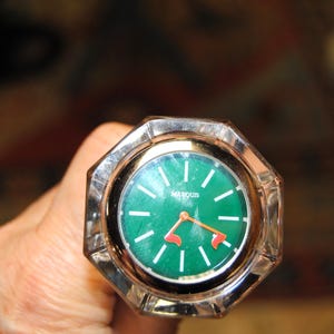 May include: A vintage watch with a green face and white hour markers. The watch has a silver-colored, octagonal case and orange hands. The brand name "MARQUIS" is printed on the watch face.