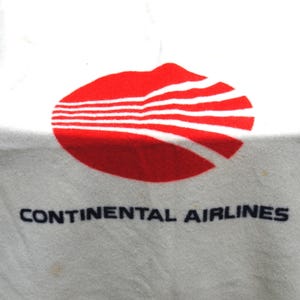 Peut inclure: Un article textile blanc arborant le logo rouge de Continental Airlines, composé d'une forme ovale traversée par cinq lignes blanches horizontales. Sous le motif rouge, le texte CONTINENTAL AIRLINES est imprimé en noir.