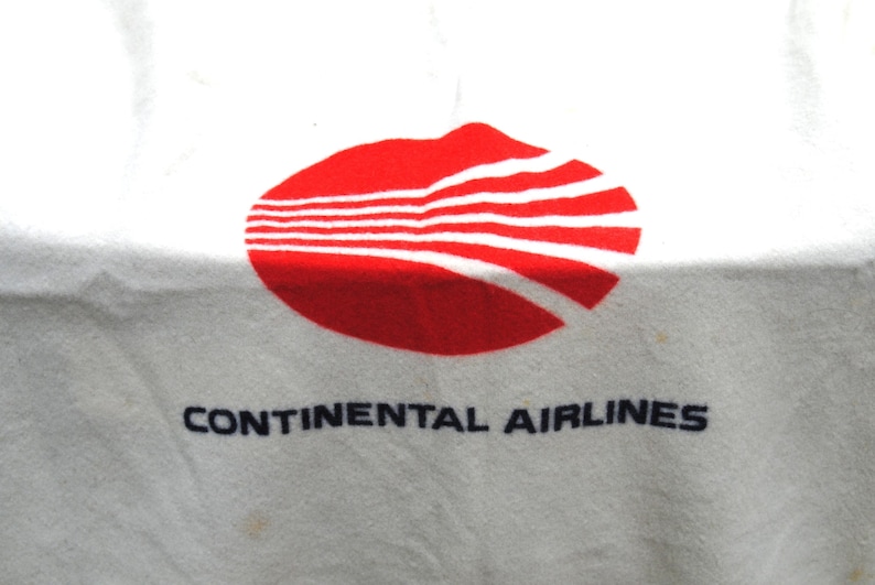 May include: A white textile item featuring the red Continental Airlines logo, which consists of an oval shape with five horizontal white lines cutting through it. Below the red graphic, the text CONTINENTAL AIRLINES is printed in black.