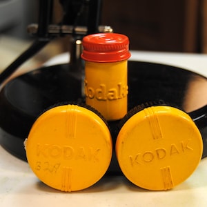May include: A collection of vintage Kodak film canisters and accessories. The image features a yellow film container with a red cap, and two yellow film container lids. The word "Kodak" is embossed on the lids and the film container.