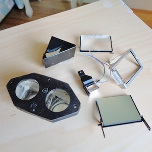 May include: A set of black and silver camera parts, including a viewfinder, a lens, and a housing with two circular openings. The parts are laid out on a light wood surface.