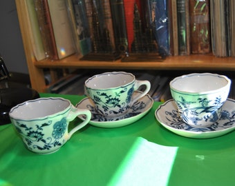 May include: Three white teacups with blue floral patterns and matching saucers. The cups and saucers are arranged on a green surface.