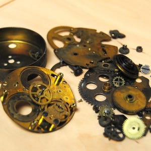 Puede incluir: Piezas de reloj antiguo desmontadas, incluyendo engranajes, placas y una carcasa. Los componentes son de varios tonos dorados, bronce y negro, lo que sugiere edad y desgaste. La disposición está dispersa sobre una superficie de madera clara.