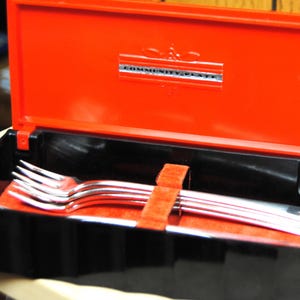 Może przedstawiać: Zestaw srebrnych widelców w czarno-czerwonym pudełku. Pudełko jest otwarte, ukazując widelce spoczywające na czerwonej filcowej wyściółce. Na pokrywie pudełka widnieje napis "COMMUNITY PLATE" w srebrnych literach.