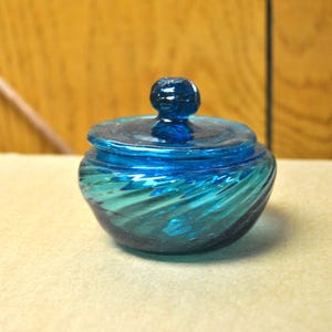 May include: A blue glass jar with a swirl design and a matching lid. The jar has a rounded shape and a smooth, glossy finish.
