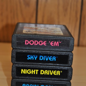 May include: A stack of four black Atari video game cartridges. The titles are "DODGE 'EM" in pink, "SKY DIVER" in blue, "NIGHT DRIVER" in yellow, and "BRAIN GAMES" in blue. The cartridges are stacked on top of each other.