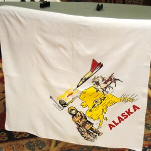 May include: White square fabric with a colorful illustration of Alaska. The design includes a yellow map of Alaska, a red and yellow airplane, a person in traditional clothing, and the word "ALASKA" in red.