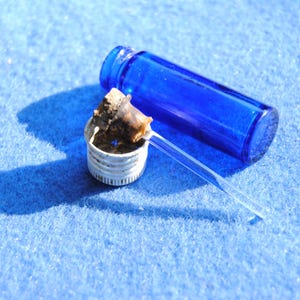 以下が含まれることがあります： 水平に置かれた小さな円筒形のコバルトブルーのガラス瓶。その隣には、白い金属製のスクリューキャップがあり、キャップの中央から伸びる細い透明なガラス製アプリケーターロッドにコルク栓が取り付けられています。