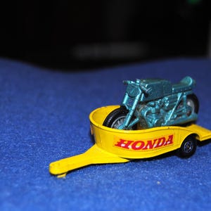 May include: A die-cast metal toy motorcycle in a metallic teal finish, secured inside a bright yellow trailer. The trailer features the word HONDA printed in bold red capital letters on its side, with black wheels and a hitch attachment.