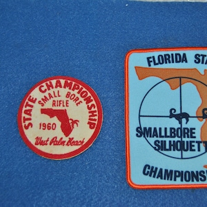 May include: Two vintage patches on a blue background. One is a red and white circular patch that reads "State Championship Small Bore Rifle 1960 West Palm Beach". The other is a light blue patch with the text "Florida State Smallbore Rifle Silhouette Championships".