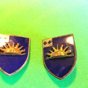 Pode incluir: Dois broches vintage em forma de escudo. Cada broche apresenta um fundo de esmalte azul com um desenho de raios de sol dourados. Um tem uma seção superior branca e dourada, o outro tem um desenho de cruz dourada.
