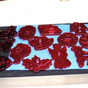 May include: A collection of red cookie cutters in various shapes, including an angel, a lamb, a butterfly, and a maple leaf. The cutters are arranged on a blue felt surface.