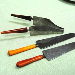 May include: A set of four metal serving utensils including a pair of tongs with dark brown handles and two flat spatulas with orange handles. The metal blades are silver-toned and have a smooth, flat surface with a tapered design for serving.