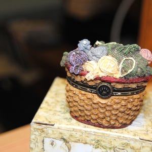 以下が含まれることがあります： カラフルな彫刻の花で満たされた、織りバスケットの形をした装飾的な小物入れ。箱は茶色で赤いトリムと黒い留め金が付いています。花は紫、黄色、緑など、さまざまな色です。