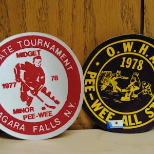 Puede incluir: Dos pegatinas redondas vintage con temática de hockey. Una es roja y blanca, con el texto "N.Y. State Tournament Midget" y "Niagara Falls N.Y.". La otra es negra y amarilla, con el texto "O.W.H.A. 1978 PEE-WEE ALL STARS".