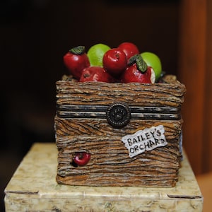May include: A decorative wooden box filled with red and green apples. The box has a rustic, textured appearance with a metal clasp and the text "Bailey's Orchard" on a small tag.